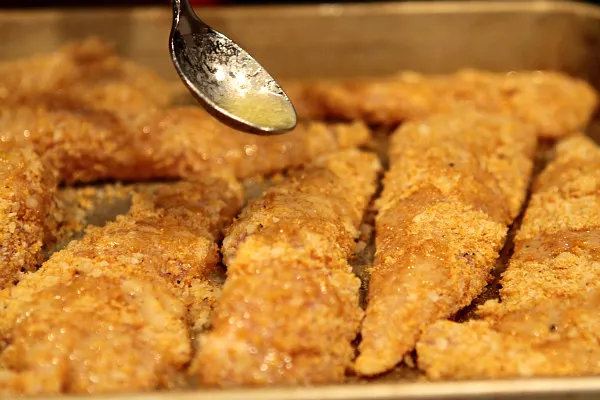 spooning butter onto parmesan chicken tenders on a baking sheet