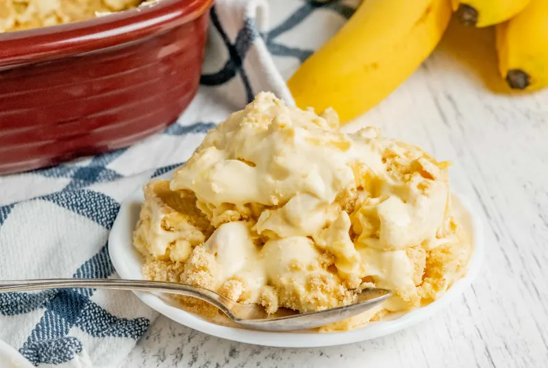 serving of banana pudding on a white plate on a blue plaid napkin with bananas and a serving dish in the background