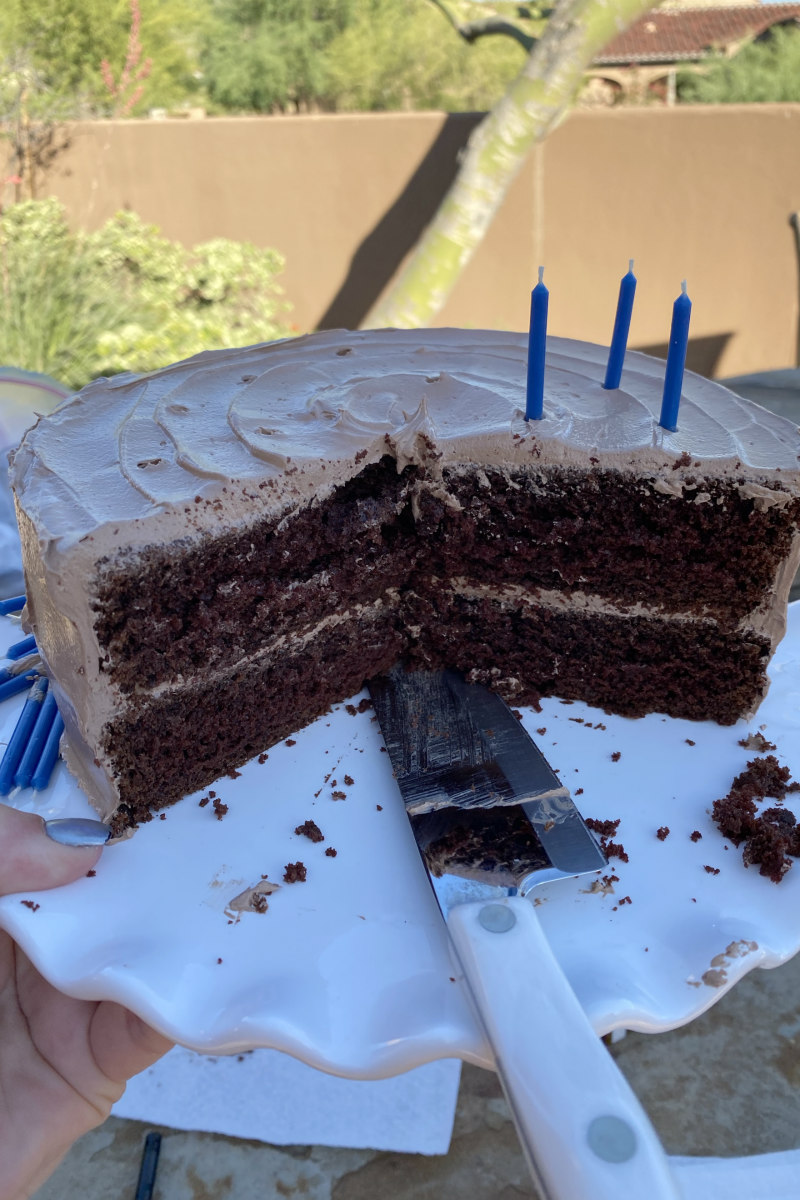 Chocolate Cake on a white platter sliced open to see the inside. Sharp knife sitting on the cake plate. Candles on top. Backyard scene.