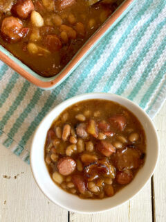 single serving of barbecue beans in a white bowl with a casserole dish filled with barbecue beans alongside, set on a blue and white striped dishtowel