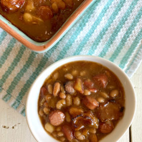 single serving of barbecue beans in a white bowl with a casserole dish filled with barbecue beans alongside, set on a blue and white striped dishtowel