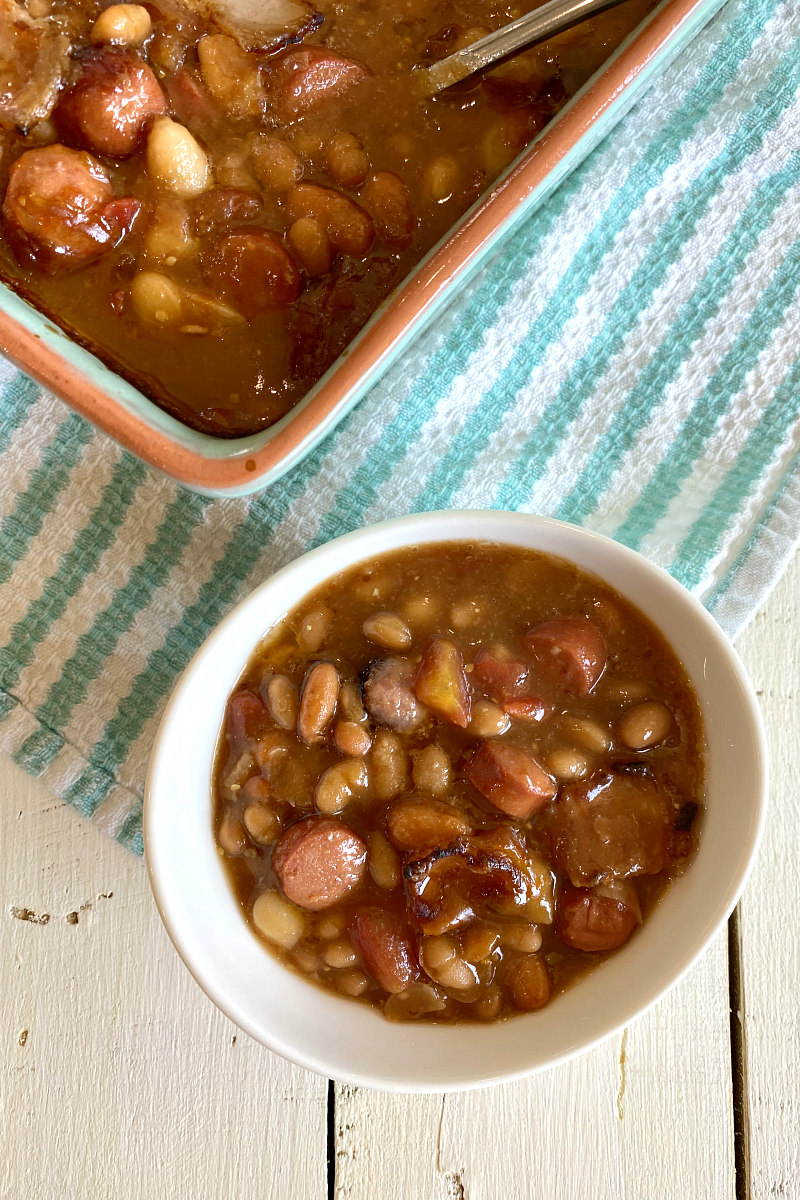 single serving of barbecue beans in a white bowl with a casserole dish filled with barbecue beans alongside, set on a blue and white striped dishtowel