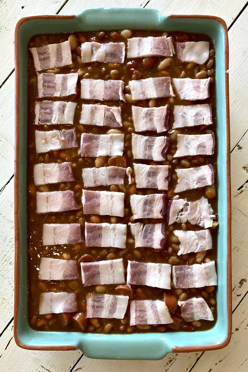 blue casserole dish filled with barbecue beans and topped with raw pieces of bacon ready for the oven