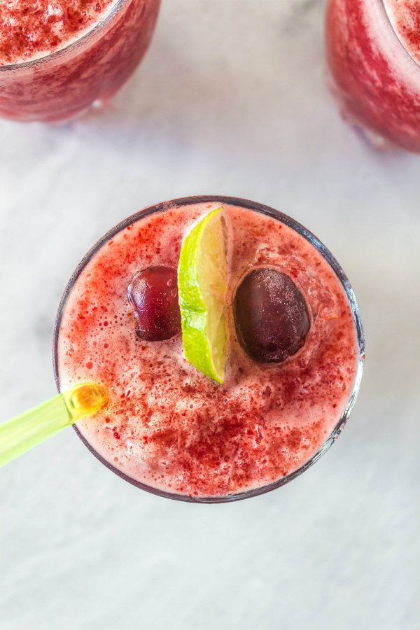 overhead shot of cherry lime slushie garnished with fresh lime and fresh cherries. peek at two more slushies in the background