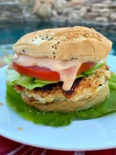 chicken and sun dried tomato burger dressed with tomato, lettuce and special sauce sitting on a bed of lettuce on a white plate set on a red plaid napkin with a backyard pool background