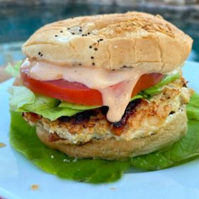 chicken and sun dried tomato burger dressed with tomato, lettuce and special sauce sitting on a bed of lettuce on a white plate set on a red plaid napkin with a backyard pool background