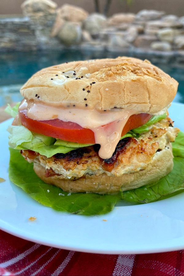 chicken and sun dried tomato burger dressed with tomato, lettuce and special sauce sitting on a bed of lettuce on a white plate set on a red plaid napkin with a backyard pool background