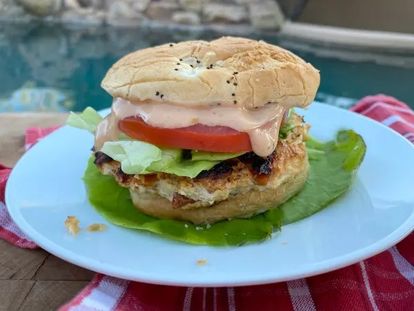 chicken and sun dried tomato burger dressed with tomato, lettuce and special sauce sitting on a bed of lettuce on a white plate set on a red plaid napkin with a backyard pool background