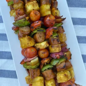 hawaiian chicken sausage kabobs stacked on a white platter set on a white/blue striped napkin
