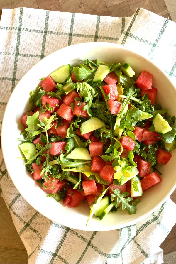 white bowl of watermelon cucumber and feta arugula salad displayed on a white and green plaid napkin