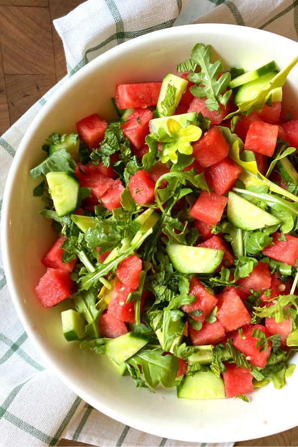 white bowl of watermelon cucumber and feta arugula salad displayed on a white and green plaid napkin