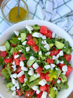 white bowl of watermelon cucumber and feta arugula salad displayed on a white and green plaid napkin