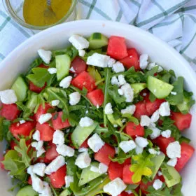 white bowl of watermelon cucumber and feta arugula salad displayed on a white and green plaid napkin