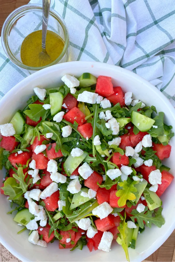 white bowl of watermelon cucumber and feta arugula salad displayed on a white and green plaid napkin