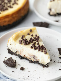 slice of chocolate chip cookie dough cheesecake on a white plate with a peek at the cheesecake in the background