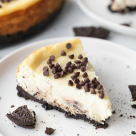slice of chocolate chip cookie dough cheesecake on a white plate with a peek at the cheesecake in the background