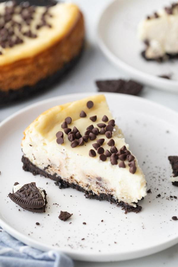 slice of chocolate chip cookie dough cheesecake on a white plate with a peek at the cheesecake in the background