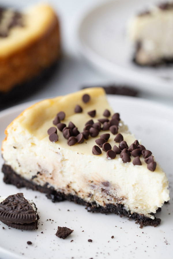 slice of chocolate chip cookie dough cheesecake on a white plate with a peek at the cheesecake in the background