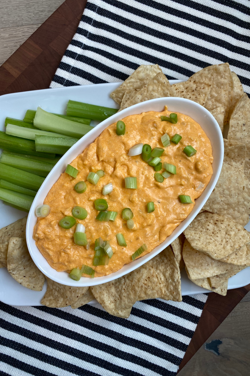Buffalo Chicken Dip in white bowl with chips and celery on black and white striped towel