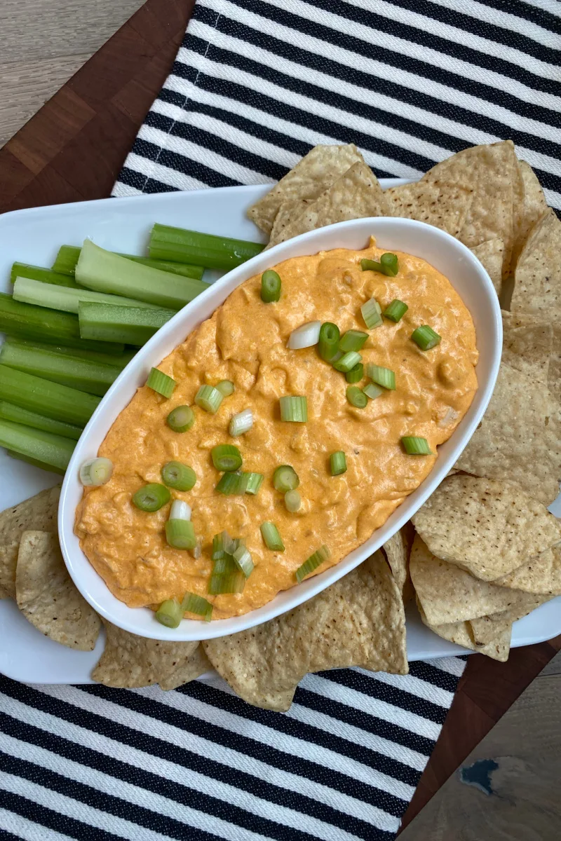 Buffalo Chicken Dip in white bowl with chips and celery on black and white striped towel