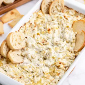 casserole dish with cheesy lemon rosemary artichoke dip and baguette