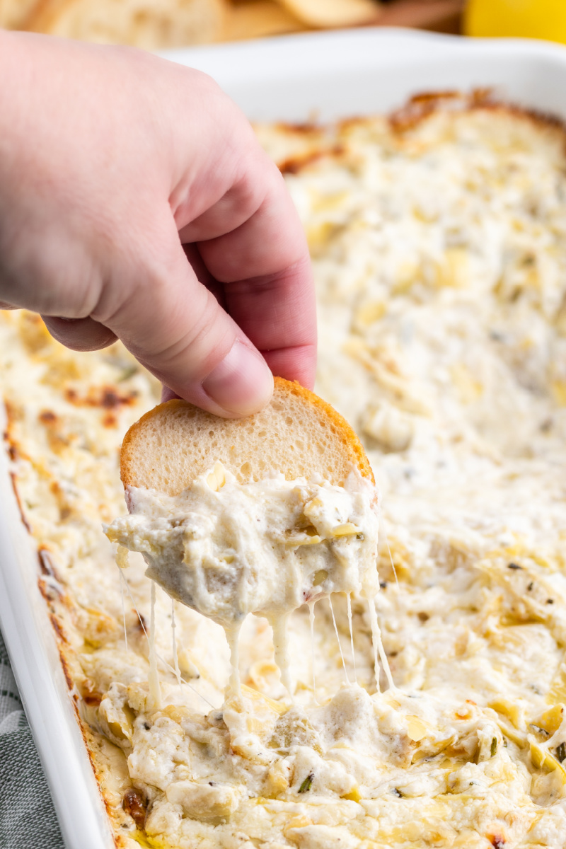 hand dipping baguette into cheesy lemon rosemary artichoke dip