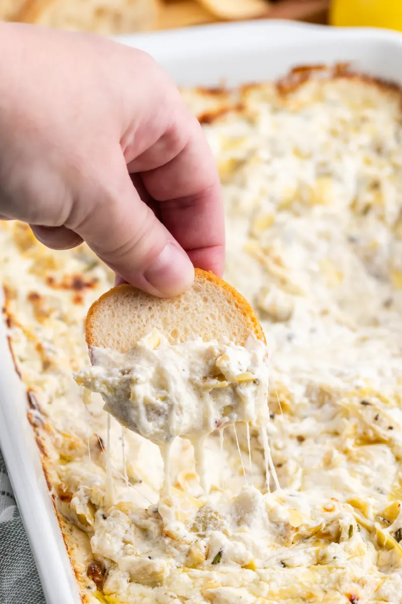 hand dipping baguette into cheesy lemon rosemary artichoke dip