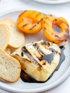 chunk of melted brie on plate with balsamic glaze, baguette and grilled apricots