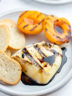 chunk of melted brie on plate with balsamic glaze, baguette and grilled apricots